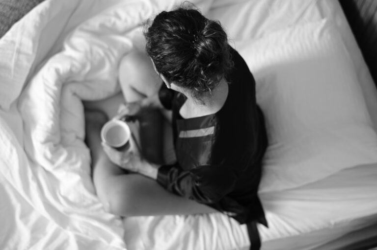 Kvinna med kaffekopp sitter i säng, ovanifrån i svartvitt