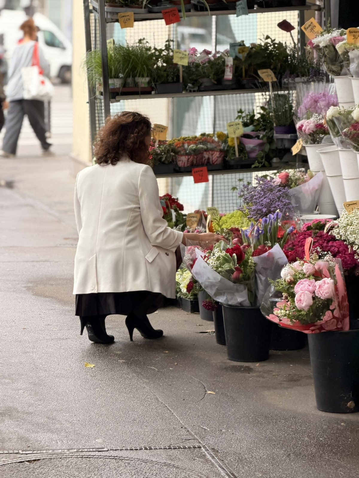 Klara i ljus kappa sitter på huk vid ett blomsterstånd på gatan och väljer blommor
