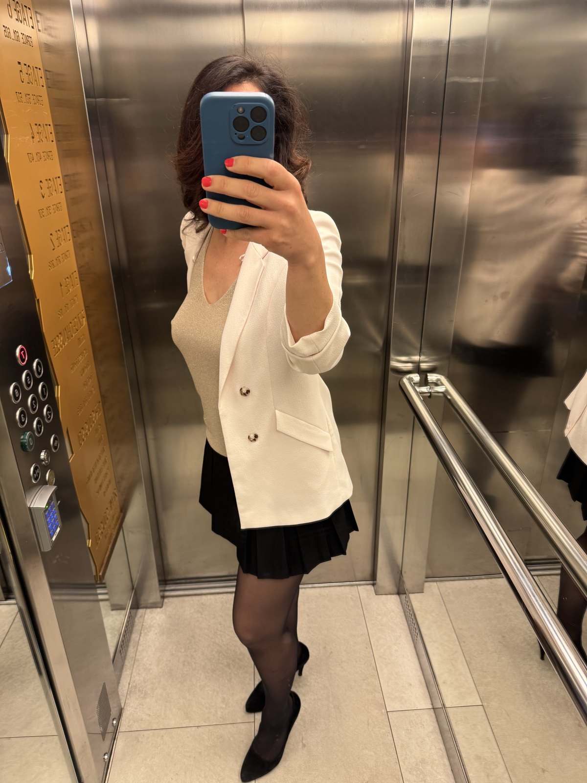Klara tar en spegelselfie i hissen, vit kavaj och svart plisserad kjol, mobilen döljer ansiktet